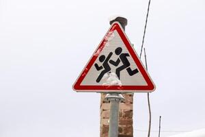Road sign depicting running children, covered with snow against a white winter sky with a chimney behind it photo