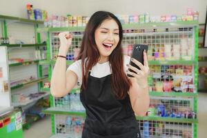 emocionado asiático mujer como un cajero es vistiendo negro delantal y participación su teléfono pantalla foto