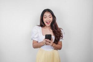 A cheerful Asian woman is smiling and holding her smartphone, isolated by white background. photo