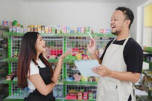 sonriente asiático personas participación un tableta, cajero es vistiendo negro y crema delantal en pie en un comestibles o conveniente Tienda foto
