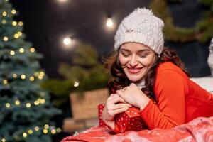 contento hermosa mujer participación Navidad presente con su ojos cerrado. dama en rojo ropa y blanco gorra acuerdo presente a su corazón. bonito niña a Navidad decoración antecedentes. foto