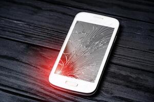 Modern smartphone with large broken screen with debris. Damaged expensive device on the dark wooden surface with light leaks. Close-up photo