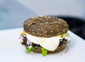 Appetizing black bun burger with fried egg. Brown bread for hamburger with sesame seeds on white background. Delicious fast food close-up. photo