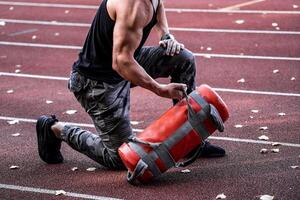 fuerte masculino haciendo rutina de ejercicio exterior. levantamiento pesado 10 kilo herramienta. hermoso deportista. joven carrocero. yendo deporte. campo deportivo antecedentes. de cerca foto