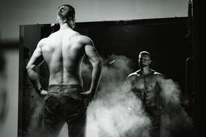 An athletic man standing and posing in front of the mirror. View from the back. Black and white photo. Smoke and power. Fitness concept. photo