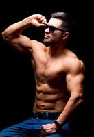 Well build handsome man without a shirt posing in studio. Strong body and cool face. Male wearing glasses, sitting on a chair with hands on legs. Closeup. photo