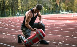 fuerte masculino haciendo rutina de ejercicio exterior. levantamiento pesado 10 kilo herramienta. hermoso deportista. joven carrocero. yendo deporte. campo deportivo antecedentes. de cerca foto