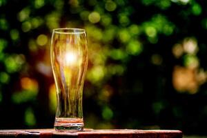 Close-up picture of an ampty beer glass on a brown wooden table. Sun shines through the glass. Blurred green nature background. photo
