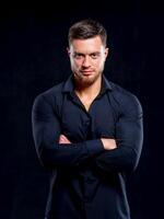 Young man in black shirt smiling isolated on dark background. Standing cross hands. Muscular male, athletic. Fashion portrait photo