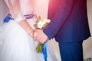 hermosa joven Pareja en pie cerca a uno otro durante el Boda ceremonia. novia en blanco vestir con azul cinturón y novio vistiendo elegante traje participación manos juntos. foto