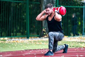 Handsome young athlete working out on a running track. Young man doing squats with a heavy bag on the track. Sport and wellbeing concept. Closeup photo