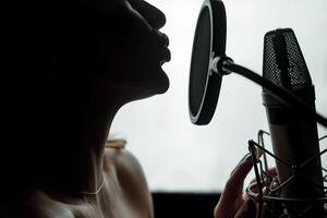 Dark silhouette profile of a young female with bare shoulders singing at the microphone isolated in studio. Face of a sexy woman performing music close to the mic. Close-up photo