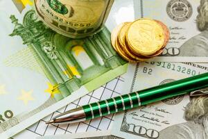 green pen lays on dollar bills near the gold coins on the table. Close-up photo