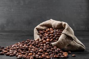 Bag with scattered coffee beans isolated on the black background. Coffee beans lying on the dark surface falling out the sack. Close-up photo