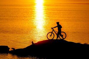 silueta de deportista montando un bicicleta en el playa. vistoso puesta de sol nublado cielo en antecedentes foto