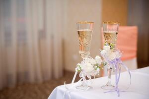 Romantic view of wedding glasses with champagne on white tablecloth in the restaurant. Two crystal glasses with sparkling champagne decorated with white flowers. Close-up photo