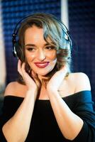 Portrait of beautiful joyful woman with bare shoulders listening to music on headphones in studio. Happy pretty female with headphones looking side. photo