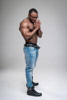 African man in good physical shape standing with his hands folded in front of his face. Strong dark-skinned male with naked torso wearing jeans only. Man standing half-turned in the white background. photo