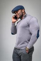 African man standing half-turned and speaking on the mobile phone. Muscular male wearing grey casual clothes and cap. Black man speaking on the phone in white background. photo