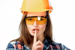 Portrait of a beautiful architect woman in safety orange helmet and protective glasses making gesture of silence by her finger. Close-up photo