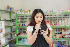 Thoughtful Asian people holding her phone, cashier is wearing black apron standing in a groceries photo