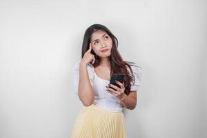 A thoughtful Asian woman is holding her smartphone while imagining her thoughts, isolated by white background. photo