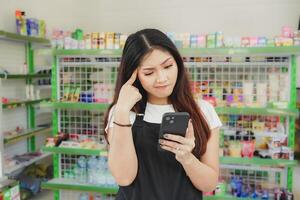 Thoughtful Asian people holding her phone, cashier is wearing black apron standing in a groceries photo