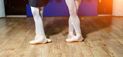el piernas de dos joven bailarinas en blanco medias y pointes ejecutando un danza en de madera piso en un estudio. joven bailarinas piernas en en pie en ballet posición. foto