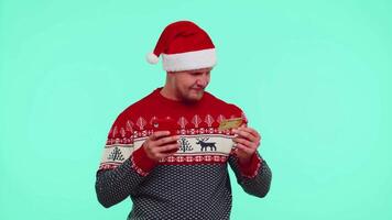 Man in Christmas sweater using credit bank card, smartphone while transferring money shopping online video