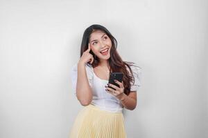 A thoughtful Asian woman is holding her smartphone while imagining her thoughts, isolated by white background. photo