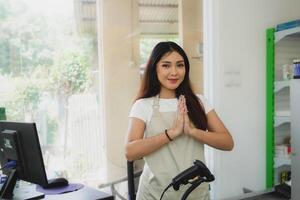 Friendly Asian female as a cashier is giving gestures of traditional greetings to customer, wearing cream apron photo