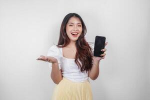 A cheerful Asian woman is holding her smartphone and pointing copy space beside her, isolated by white background. photo