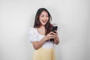 A cheerful Asian woman is smiling and holding her smartphone, isolated by white background. photo