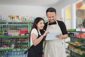 sonriente asiático personas participación un tableta, cajero es vistiendo negro y crema delantal en pie en un comestibles o conveniente Tienda foto