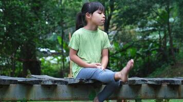 Cute little girl upset sitting on a wooden bridge near the river in summer. little girl sitting alone on a wooden bridge looking at the river outdoors. video