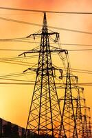 Dark silhouettes of transmission lines standing one by one in a row. Power lines against the backdrop of setting sun. Pink and yellow sky crossed by the power wires. photo