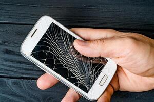 Man's hand holds mobile phone with broken touchscreen on dark background. White cracked smartphone in the male's hand. Close-up photo