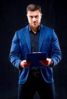 Half-length portrait of handsome young smiling man wearing dark shirt and blue suit holding blue folder, looking at camera. Black background. Money and business concept. photo