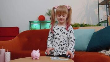 Little child kid girl putting money dollar banknotes for future gift purchase at piggybank at home video