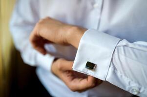 manos de Boda novio abotonarse arriba su blanco camisa. masculino manos en un antecedentes de un blanco camisa, manga camisa con gemelos. de cerca foto