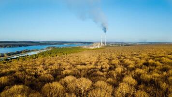 Birds-eye view of a beautiful nature in spring with a background of a factory with two big pipes. Brown forest without leaves near the river under the blue sky outdoors. photo