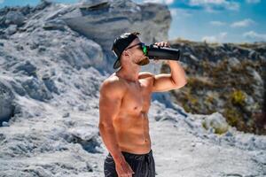 retrato de un hermoso medio desnudo carrocero hombre en un montaña con un botella de agua. foto