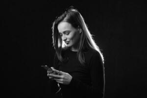 Black and white portrait of young woman looking at phone and smiling, isolated studio photo on dark background. Beautiful model with long hair using cell phone