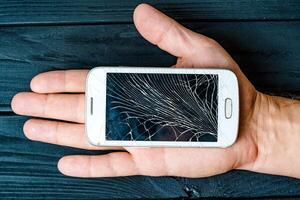 Man's hand holds mobile phone with broken touchscreen on dark background. Smartphone with cracked broken screen in man's palm. photo