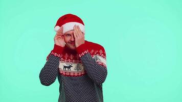 Man in sweater Santa Christmas hat fooling around having closing eyes with hand and spying through video