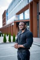 Handsome dark-skinned man standing near building. Fit man in tight shirt and glasses looking straight ahead. Man with big biceps in a tight shirt at business center. photo