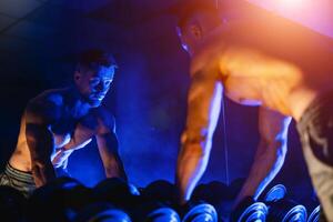 A young man watches in the mirror performing a biceps holding chrome dumbbell. Blue light filter. Selective focus photo