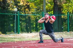 hermoso joven atleta trabajando fuera en un corriendo pista. joven hombre haciendo sentadillas con un pesado 10 kilo bolso en el pista. deporte y bienestar concepto. de cerca. foto