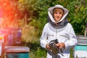 Beekeeper standing with equipment. Man in protective uniform near beehives wiht honey and bees. photo