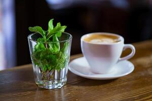 transparent glass with a fresh mint inside stands on the background of a cup with a latte in the cafe. Close-up photo